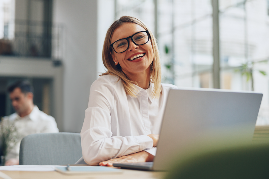 mujer sonriendo, sentada frente a un ordenador en una oficina, con personas difuminadas trabajando en segundo plano