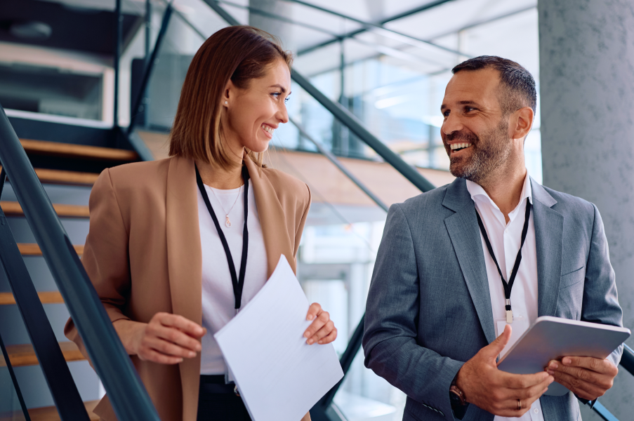Dos profesionales de negocios conversando en una oficina moderna, un hombre con tablet y una mujer con credencial, ambos sonriendo