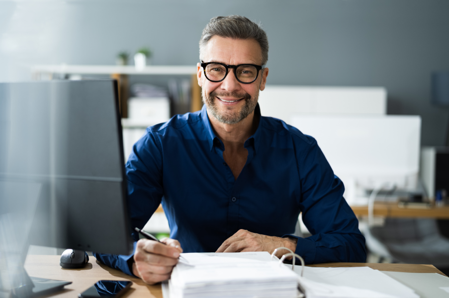 hombre sonriendo en un despacho moderno con papeles sobre su mesa y un bolígrafo en la mano
