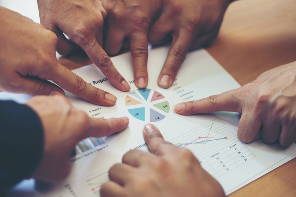 Muchas manos señalan con el dedo índice los gráficos de una hoja de informe.