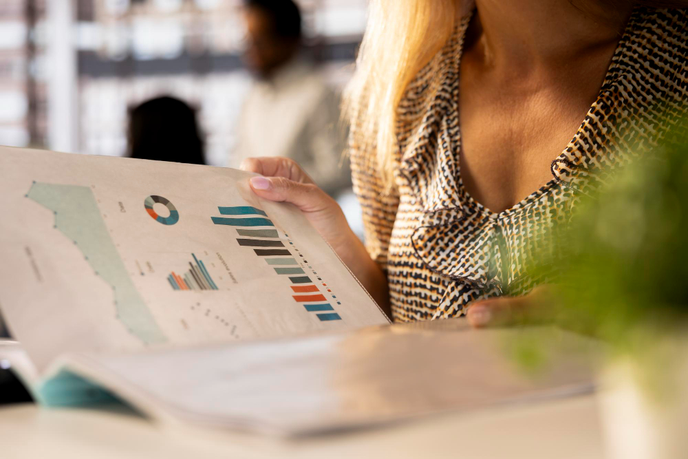 Mujer sentada en una oficina revisando un informe en papel con gráficos y datos estadísticos, en un entorno de trabajo moderno con personas desenfocadas al fondo.
