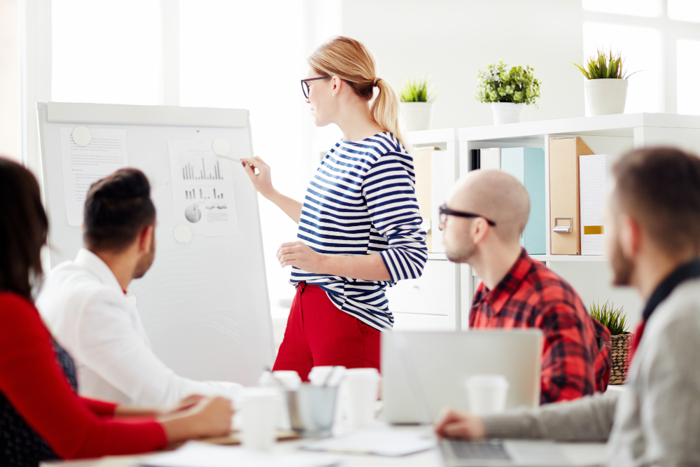 Woman presenting graphs on a whiteboard during a team meeting in an office, while several colleagues listen attentively.