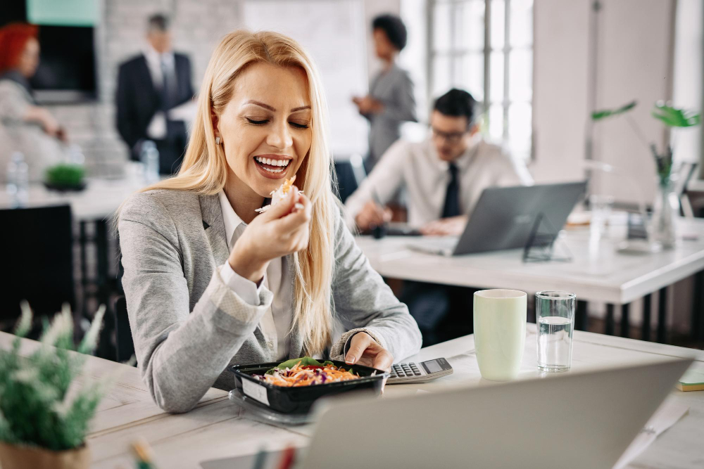Mujer sonriente almorzando en la oficina frente al portátil mientras trabaja con compañeros detrás.