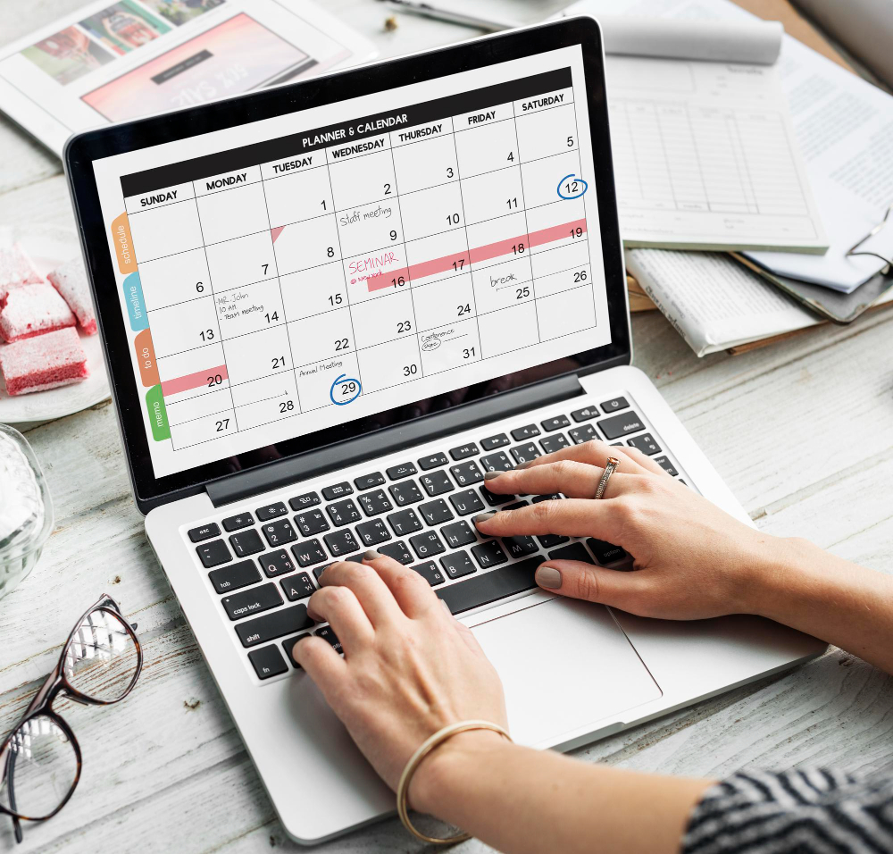 Laptop with an open digital calendar and hands organising working days and holidays in an office.